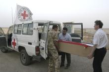 Three adult men carry a casket out of an ICRC vehicle, across a plain landscape.